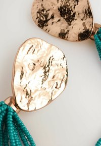 Metallische Roségold-Anhänger mit einer gehämmerten Textur, verbunden durch Stränge kleiner Türkisperlen. Glatte, glänzende Oberfläche und unregelmäßige Form.