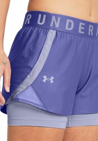 Shorts sportivi di un leggero colore viola con una vita grigia su cui è presente il testo "UNDER ARMOUR", design a strati e logo sul lato. Tessuto morbido e con texture.