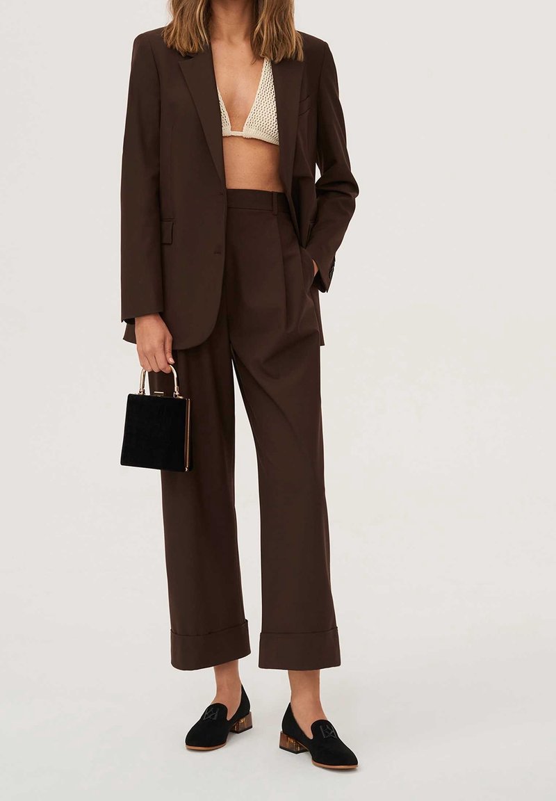 Femme portant un costume marron foncé avec un pantalon à jambes larges, un bralette crème en crochet, des mocassins noirs à talons blocs, et tenant un petit sac à main noir.