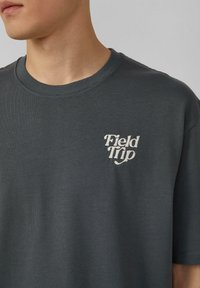 Man in een donkergrijze ronde hals T-shirt met witte geborduurde tekst "Field Trip" op de linkerkant van de borst tegen een effen achtergrond.