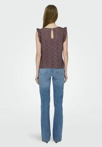 Top viola con maniche a sbuffo, con dettaglio a forma di chiave sul retro, indossato con jeans a zampa blu, che mostrano tagli floreali e un materiale testurizzato.