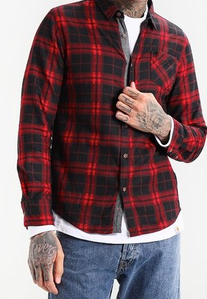 Mężczyzna w czerwono-czarnej koszuli w kratę na szarej koszulce i białym t-shircie, z widocznymi tatuażami na rękach i niebieskich dżinsach.