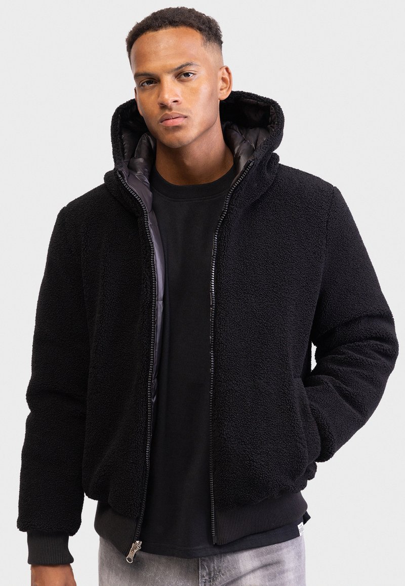 Young man wearing a black zip-up fleece jacket with hood, black shirt, and light gray jeans, standing against a plain background.
