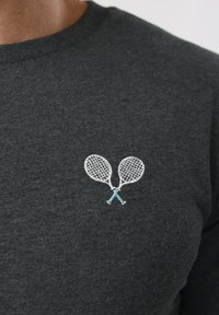 Σκούρο γκρι πουκάμισο με κεντημένα λευκά τενιστικά ρακέτες σε σχήμα σταυρού με ανοιχτό μπλε λαβές στο στήθος.