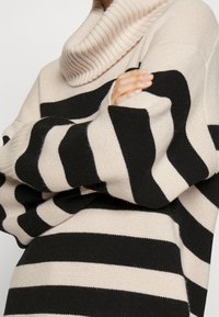 Personne portant un pull à col roulé beige et noir à larges rayures, manches longues, bras croisés, ongles soigneusement manucurés.