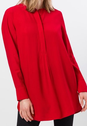 Vrouw draagt een felrode tuniek met lange mouwen en verborgen knopen aan de voorkant en zwarte geruite broek tegen een witte achtergrond.