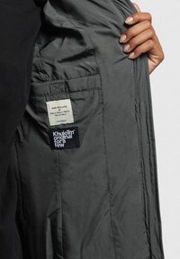 Intérieur d'une veste grise arborant une étiquette avec la mention "FAIT AVEC AMOUR PAR VRAIMENT TRÈS BEAU" et un patch de logo pour "Khujo". Texture lisse.