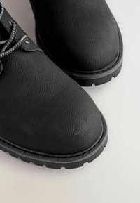 Schwarze, strukturiertes Lederstiefel mit abgerundeten Zehen und dicken, robusten Sohlen auf einem hellen Hintergrund.
