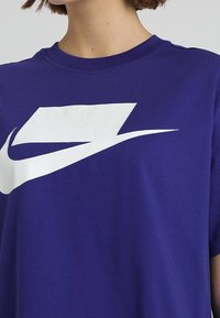 Osoba nosząca niebieski t-shirt z dużym białym abstrakcyjnym logo w kształcie zakrzywionej linii na piersi.