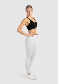 Czarny krótki top ze zmarszczonym detalem, zestawiony z białymi legginsami o wysokim stanie. Modelka w białych butach sportowych stoi w profilu.