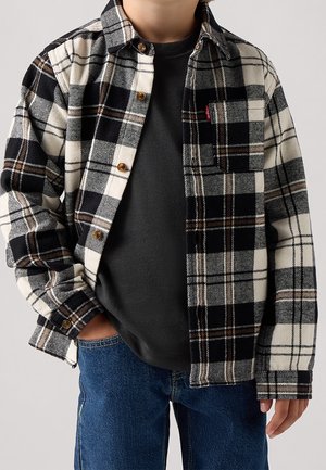 Barn iført sort-hvid ternet skjorte over mørkegrå t-shirt og blå jeans med den ene hånd i lommen.