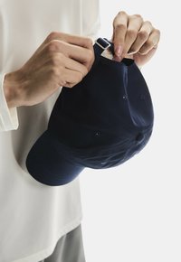 Navy baseball cap with a curved brim, fabric texture, adjustable strap, and ventilation holes, held at the opening by a hand.