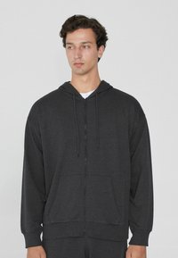 Sudadera con cremallera de color gris carbón, hecha de tela suave, que cuenta con capucha ajustable, bolsillos frontales y puños acanalados, con un corte relajado.