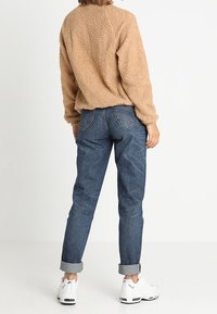 Pull en polaire marron et duveteux à manches longues, associé à un jean bleu retroussé aux chevilles et des baskets blanches. Tenue décontractée avec une finition texturée.