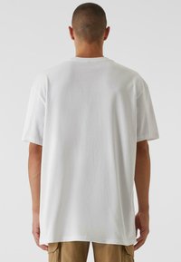 Weißes Baumwoll-T-Shirt mit lockerer Passform, kurzen Ärmeln und rundem Ausschnitt. Der Stoff wirkt glatt und untexturiert, mit einer sauberen Rückansicht.