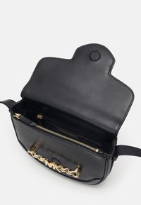 Sac à main en cuir noir avec un logo en métal doré à l'avant, doté d'une fermeture éclair et de plusieurs compartiments intérieurs.