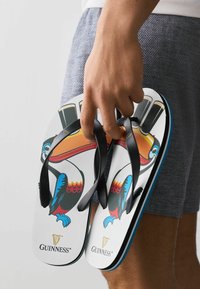 Main tenant des tongs blanches à lanières noires, avec un motif coloré de toucan et des logos Guinness, sur un fond de short gris.