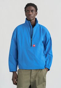 Μπλε ζακέτα με ψηλό γιακά, μπροστινό φερμουάρ και ελαστικές μανσέτες. Διαθέτει μικρό κόκκινο λογότυπο στο στήθος. Φοράται πάνω από χακί παντελόνια cargo.