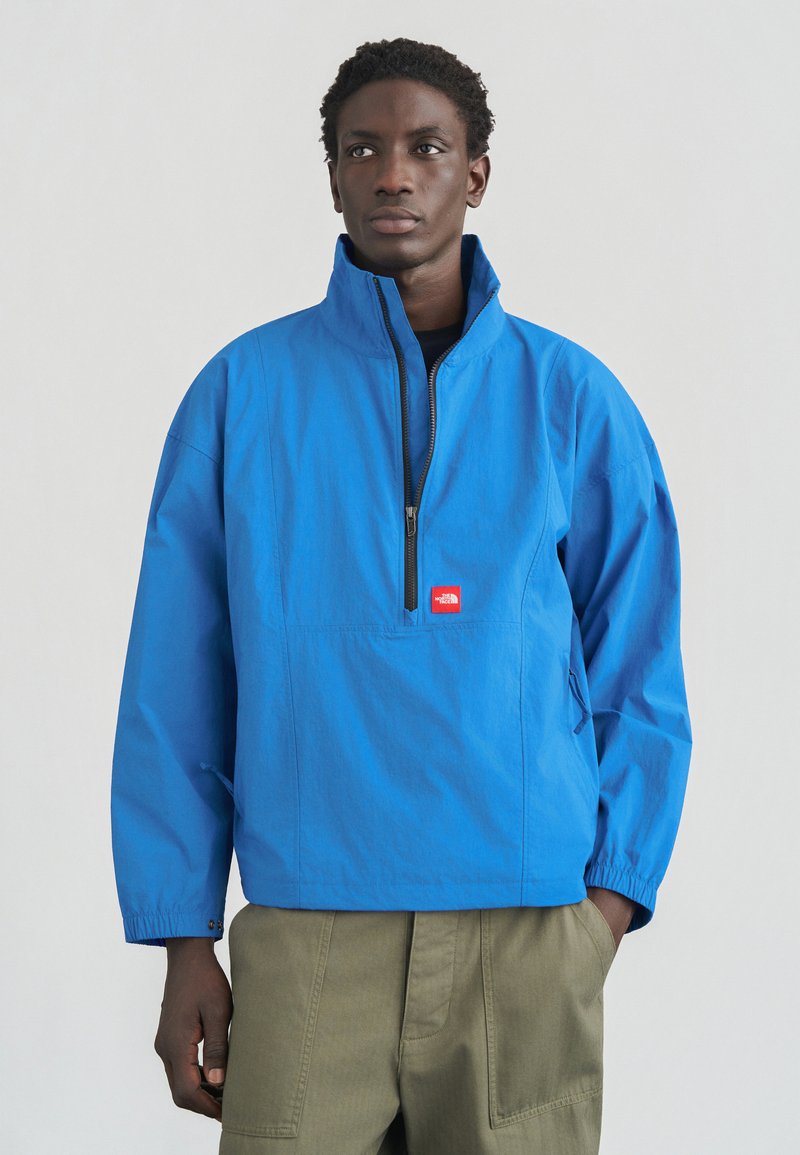 Μπλε ζακέτα με ψηλό γιακά, μπροστινό φερμουάρ και ελαστικές μανσέτες. Διαθέτει μικρό κόκκινο λογότυπο στο στήθος. Φοράται πάνω από χακί παντελόνια cargo.