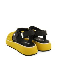 Sandalias negras con suelas de plataforma amarillas, que presentan un diseño acanalado, correas ajustables y acentos metálicos. Materiales de textura suave utilizados.