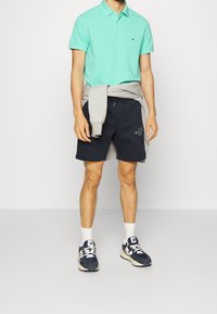 Ljus turkos polo med krage, grå sweatshirt knuten runt midjan, marinblå shorts med logotyp och marinblå sneakers med vita detaljer.