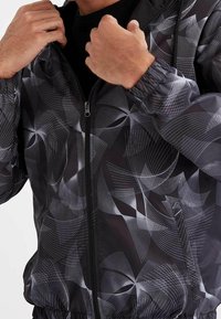 Sort vindjakke med et geometrisk hvidt linjemønster, der har lynlås, elastiske manchetter og et let materiale.