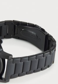 Bracelet de montre en acier inoxydable noir avec une finition mate, présentant un design segmenté, un fermoir sécurisé et une marque gravée sur la partie inférieure.