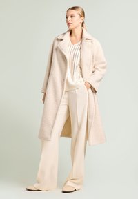 Abrigo largo texturizado color beige con solapas anchas, superpuesto sobre un top de punto crema, combinado con pantalones de pierna ancha en beige claro y zapatos claros.