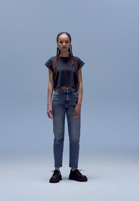 Svart cropped t-skjorte, lyseblå jeans med høy midje og svarte platåsko. Modellen står mot en lys blå bakgrunn.