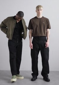 Dos modelos están juntos; uno lleva una chaqueta bomber oliva sobre un atuendo negro, el otro una camiseta marrón con pantalones y zapatos negros.