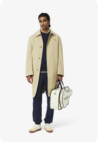 Beige regenjas met grote knopen, marineblauwe trui, marineblauwe broek en beige sneakers. Houdt een witte, gitaarvormige Lacoste tas met zwarte accenten vast.