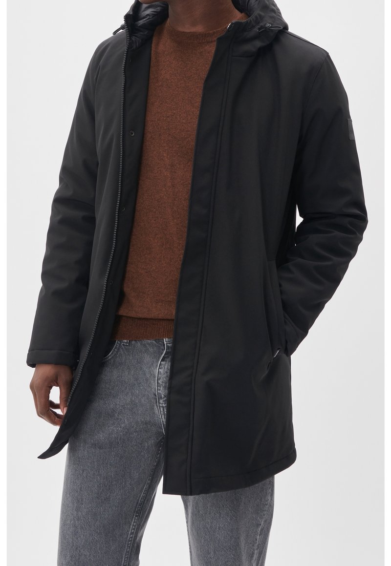 Parka noire à capuche avec fermeture éclair, dotée d'une texture lisse et de poches latérales, portée par-dessus un pull marron et un jean gris.