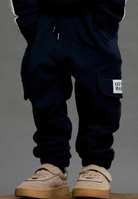 Tumesinine dressipüksid, mille pahkluude juures on elastsed cuffs, reguleeritav string ja küljetaskud. Parema jala peal on valge "LET'S PLAY" teip.