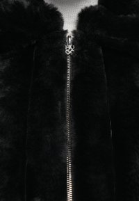 Sort faux fur jakke med en skinnende sølv lynlås, "HUGO" logo trækstang og blød tekstur, med høj krave og ingen synlige mønstre.