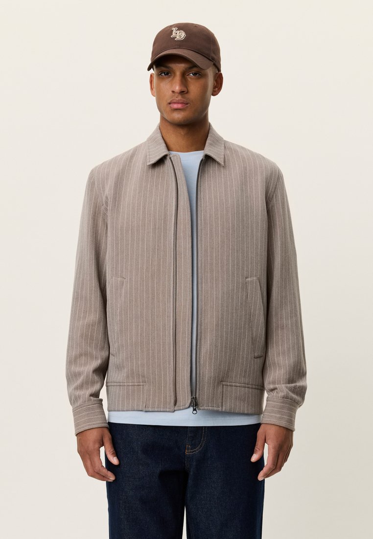 Ανοιχτό καφέ ριγέ μπουφάν με γιακά και φερμουάρ μπροστά, συνδυασμένο με μπλε πουκάμισο και σκούρο τζιν. Κατασκευασμένο από υφασμάτινη υφή.