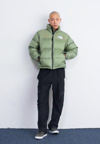 Giacca puffa verde con chiusura a zip e logo The North Face, abbinata a pantaloni cargo neri. Il modello indossa scarpe slip-on grigie.