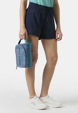 Persona con pantalones cortos azul marino de cintura elástica y zapatillas blancas, sosteniendo al lado una bolsa azul Helly Hansen con asas verdes.