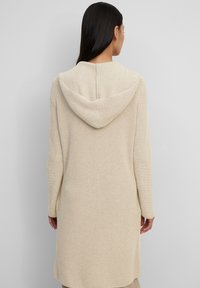 Cardigan lavorato a maglia beige di lunghezza al ginocchio, con un ampio cappuccio, maniche lunghe e un motivo testurizzato sulla superficie del capo.