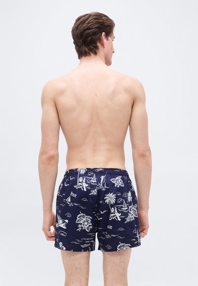 Άνδρας που βλέπεται από πίσω, φορούσε μπλε σκούρο μαγιό σορτς με λευκά τροπικά και ναυτικά σχέδια σε μονόχρωμο φόντο.