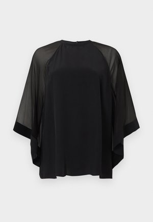 Vrouw met een zwarte top met doorschijnende mouwen, zwarte wijde broek, zwarte puntige platte schoenen en een zwarte handtas, tegen een witte effen achtergrond.