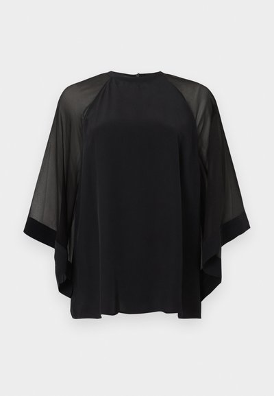 Blouse noire ample avec manches larges transparentes et encolure ronde, présentée sur un fond clair uni.