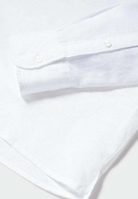 Primo piano di un polsino di una camicia bianca a maniche lunghe con due bottoni bianchi e texture del tessuto visibile su uno sfondo bianco.