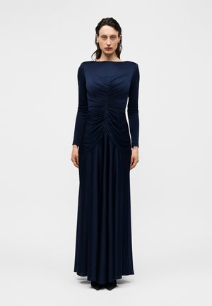 Kobieta stoi zwrócona przodem, ubrana w długą granatową sukienkę z marszczonymi detalami na górze i długimi rękawami na jednolitym tle.