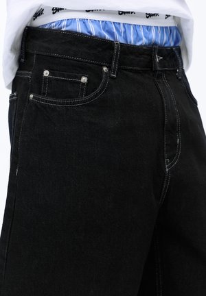 Pás čiernych džínsov z denimu s bielym prešitím, nosených cez modro-biele pruhované boxerky a bielu košeľu.