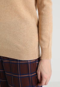 Maglione beige a maglia con polsini a coste, indossato sopra pantaloni a scacchi marroni con linee arancioni e blu. Texture morbida, design aderente.