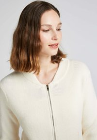 Cardigan en tricot côtelé de couleur crème avec une fermeture éclair et un col arrondi, doté de manches longues et d'une texture douce.