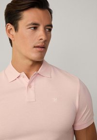 Světle růžové polo triko vyrobené z pletené tkaniny. Obsahuje límeček, dva knoflíky a malé vyšívané logo na levé straně hrudi. Krátké rukávy.