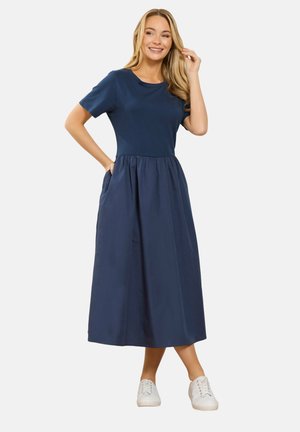 Uśmiechnięta kobieta ubrana w granatową, midi sukienkę z krótkim rękawem i kieszeniami, w połączeniu z białymi trampkami, stojąca na tle białego tła.