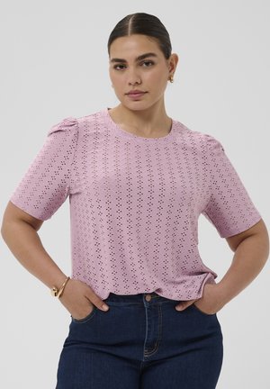 Vrouw met donker haar in een knot draagt een lichtpaars korte mouwen ooglet top en donkerblauwe jeans, staand met handen in de zakken.