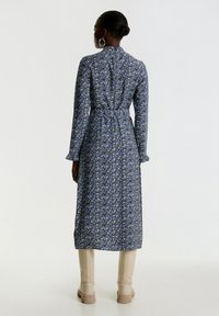 Blumenkleid in Blau und Grün, mit langen Ärmeln, in Midi-Länge, tailliert und mit fließendem Rock, kombiniert mit kniehohen beigen Stiefeln.